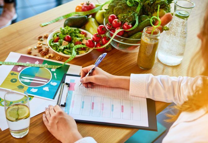 Woman dietitian in medical uniform with tape measure working on a diet plan sitting with different healthy food ingredients in the green office on background. Weight loss and right nutrition concept